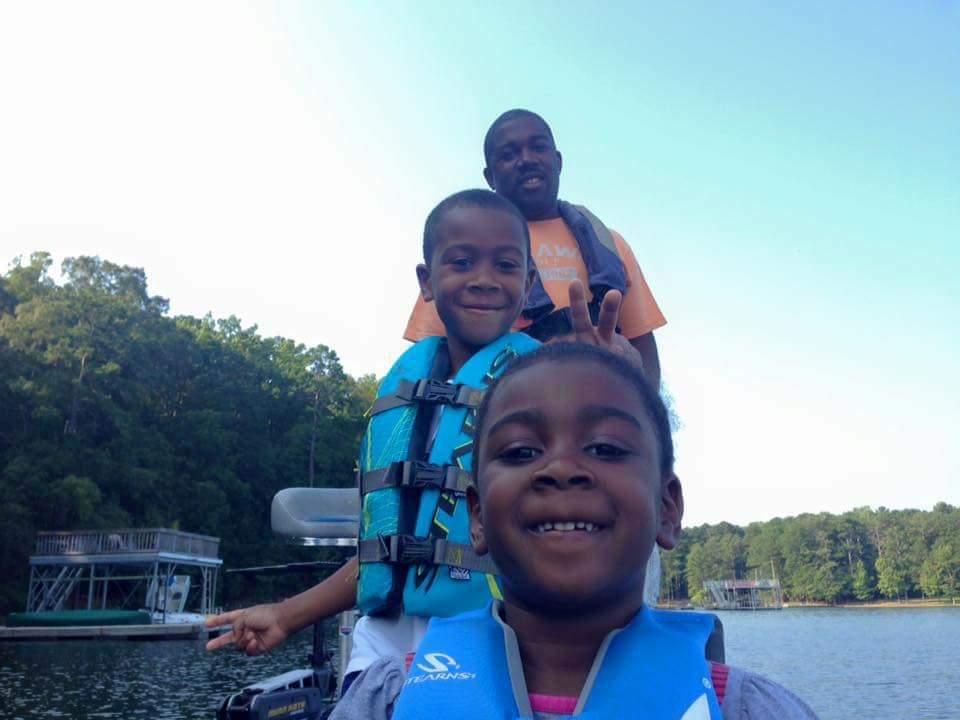 Family in life vests on a boat, smiling at the camera on a lake. Green trees in background.