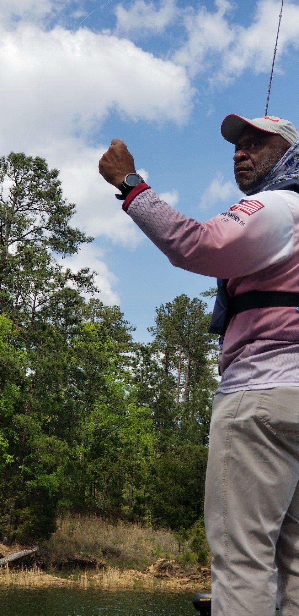 Man fishing on a boat with lush green trees and a cloudy blue sky.