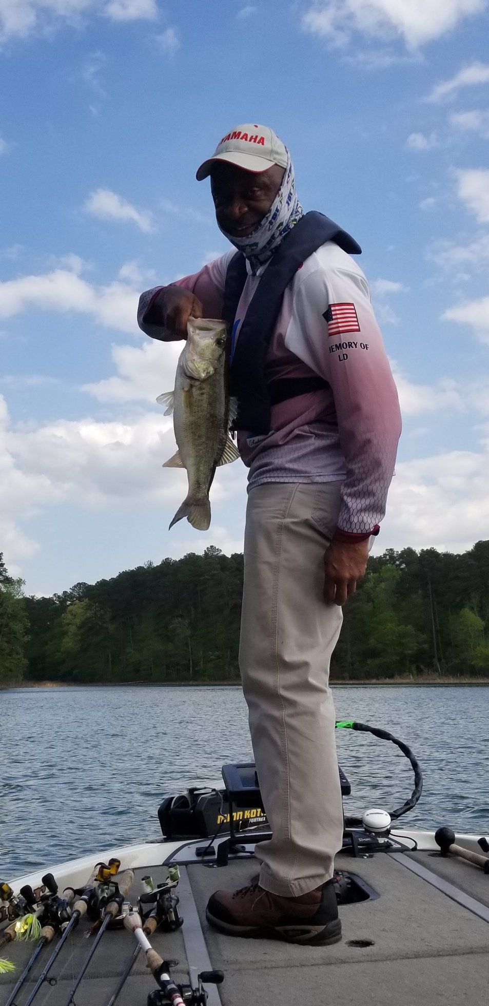 Man on a boat holds up a fish, posing for the camera with the lake and trees in the background.