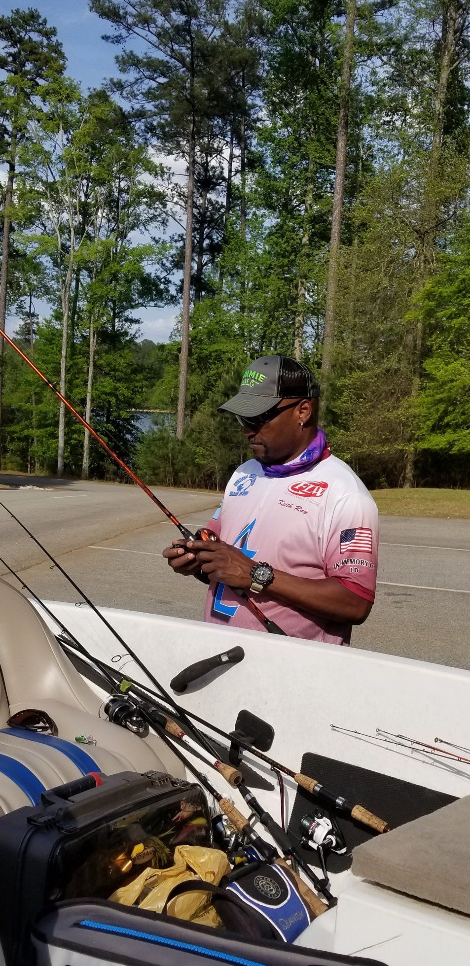 Man fishing from a boat, adjusting his rod. Green trees and blue sky in the background.