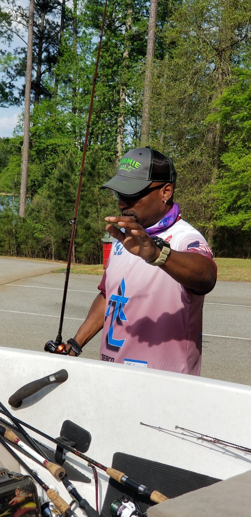 Man in fishing gear gesturing, holding a rod. Standing near a boat with fishing rods. Green trees and water in background.