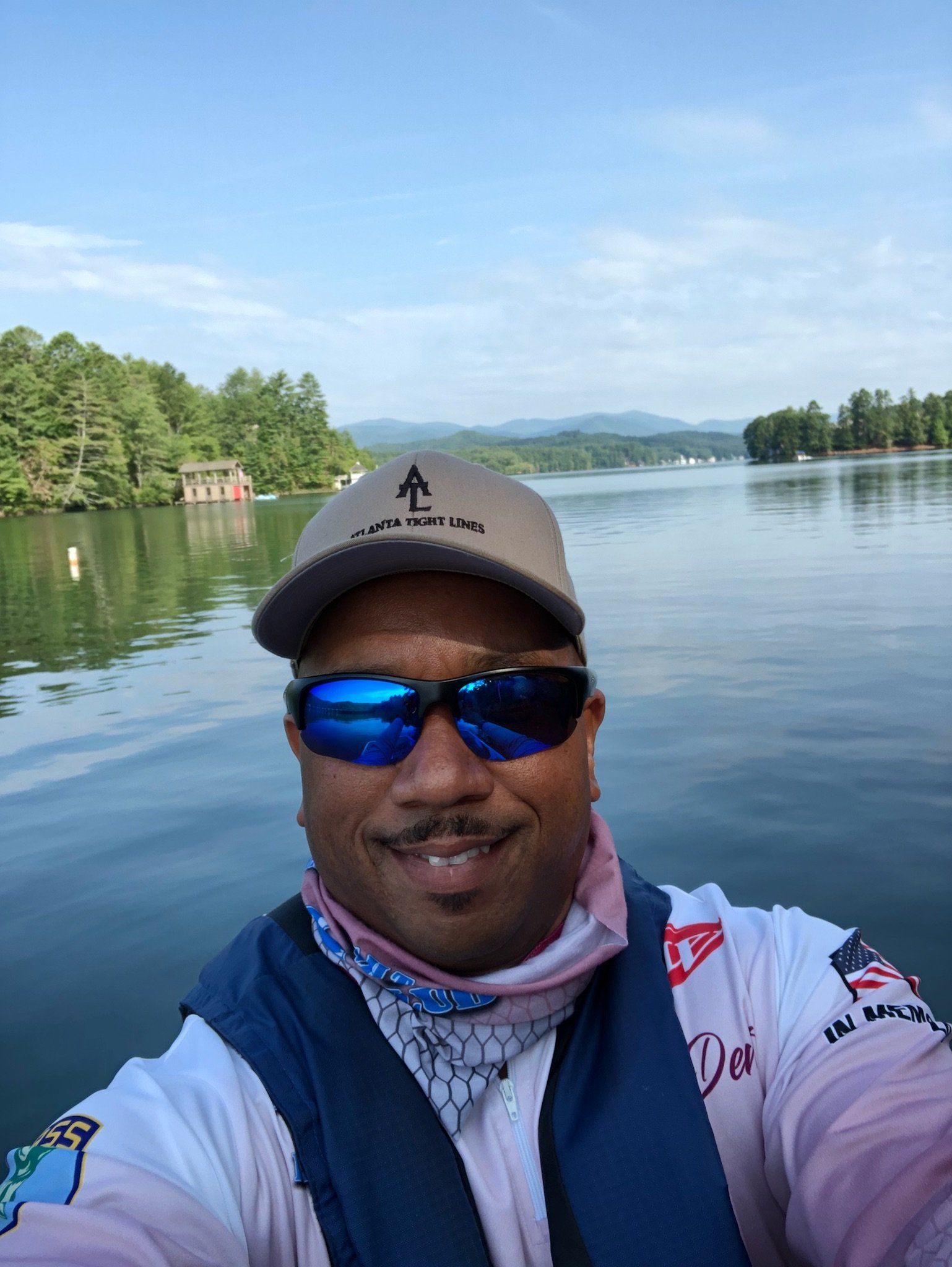 Man on boat wearing sunglasses, hat, and face covering smiles on lake with mountains in background.