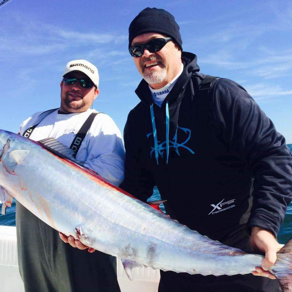 Two men on a boat holding a large, silver fish. Sunny day, blue water. Man on right smiles.