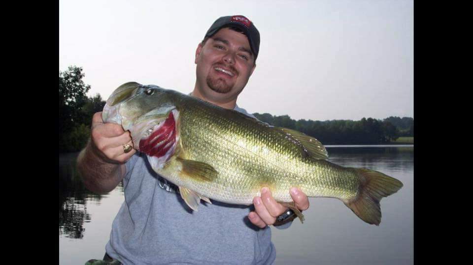 Man smiling, holding a large largemouth bass he caught near a lake.