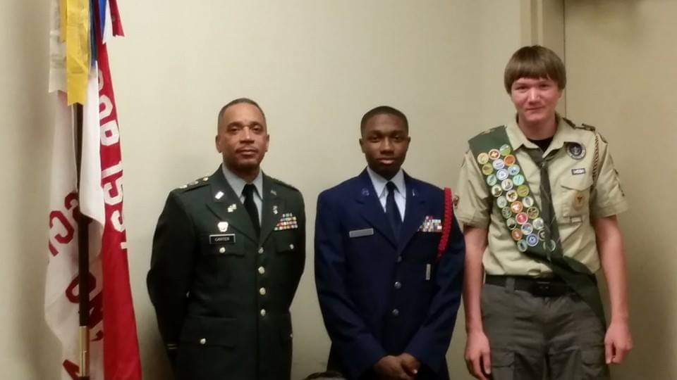 Three men in military uniforms and Boy Scout attire standing indoors. One in Army, one in Air Force, and a Boy Scout.