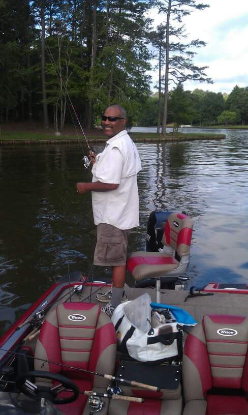 Man in a fishing boat smiling while holding a fishing rod. He wears sunglasses, tan shirt, and shorts. Trees and water in background.