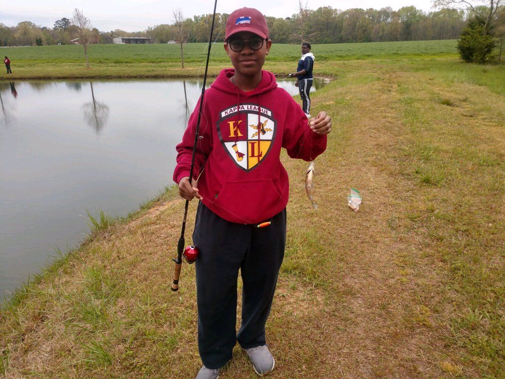 Boy in red hoodie holds up a small fish he caught by a pond.