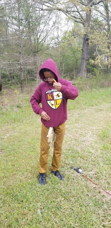 A person in a burgundy hoodie holds a fish on a grassy lawn near trees.