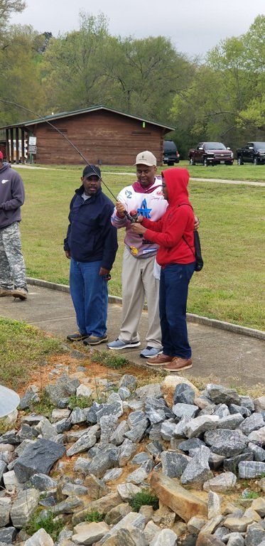 Three men by a lake, one with a fishing rod. Man in red hoodie shows something on the rod. Grassy field, rocks, and building in background.