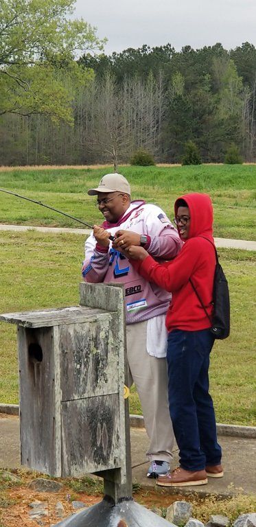 A man helps a child hold a fishing rod, near a wooden birdhouse. They are in a grassy park setting.
