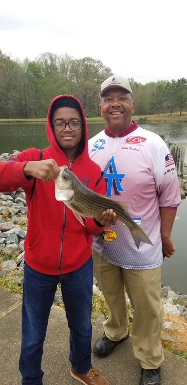 A young man in red hoodie holding a fish with an older man, near a lake. They are smiling.