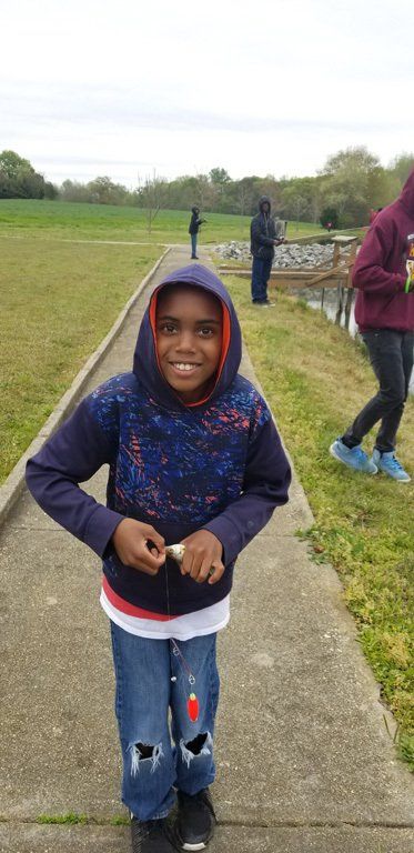 A smiling boy wearing a blue hoodie and torn jeans, holding something small, stands on a walkway.