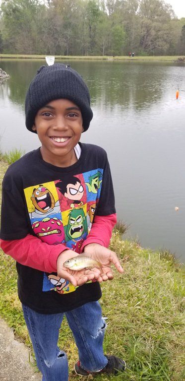 Young boy smiling, holding a small fish, standing near a lake, wearing a beanie and cartoon t-shirt.