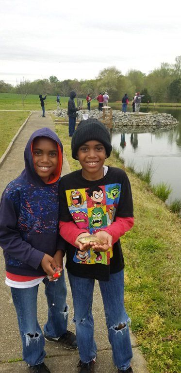 Two young boys smiling, one holding a small fish by a lake. Other people fishing in the background.
