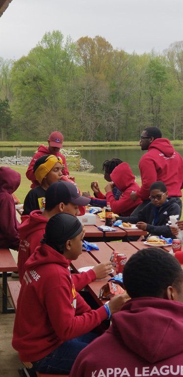 People in red Kappa League hoodies eat at a picnic table near a lake and trees.