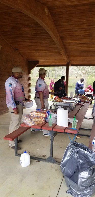 People at a picnic table under a wooden pavilion. They are prepping food and drinks.