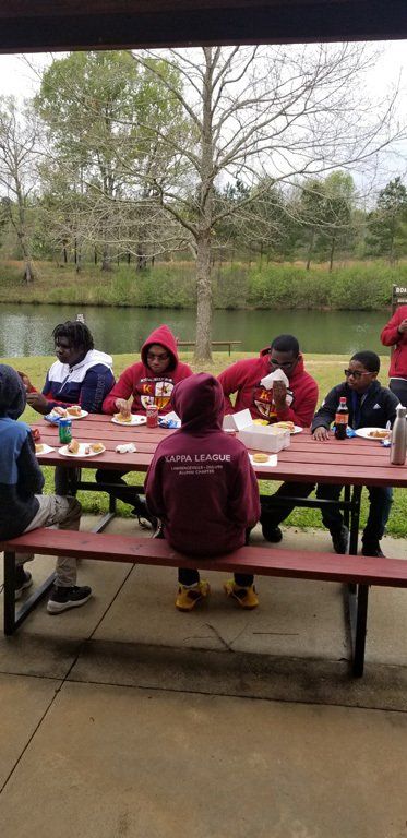 A group of people at a picnic table, eating and drinking outdoors near a body of water.