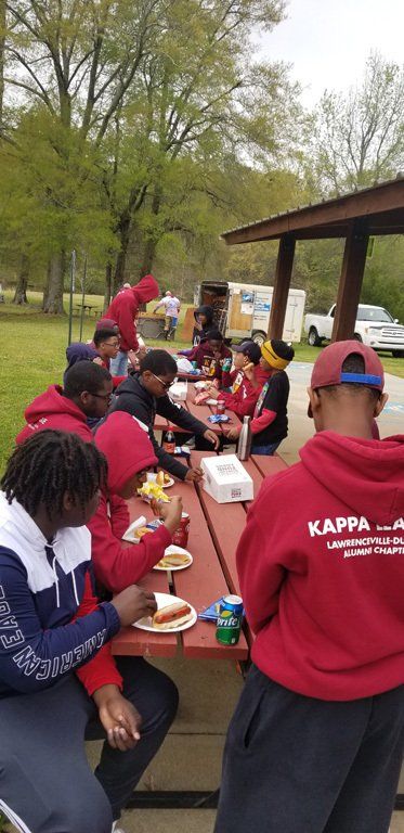Group of people at a picnic table, eating food in a park. Red and black clothing.
