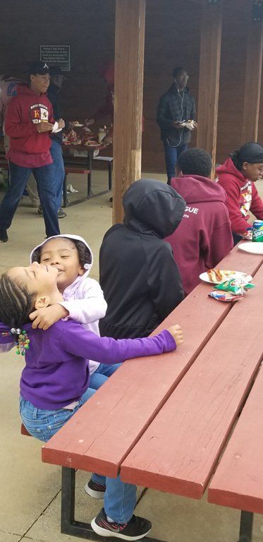 Two young girls embracing at a picnic table outdoors, other people nearby.