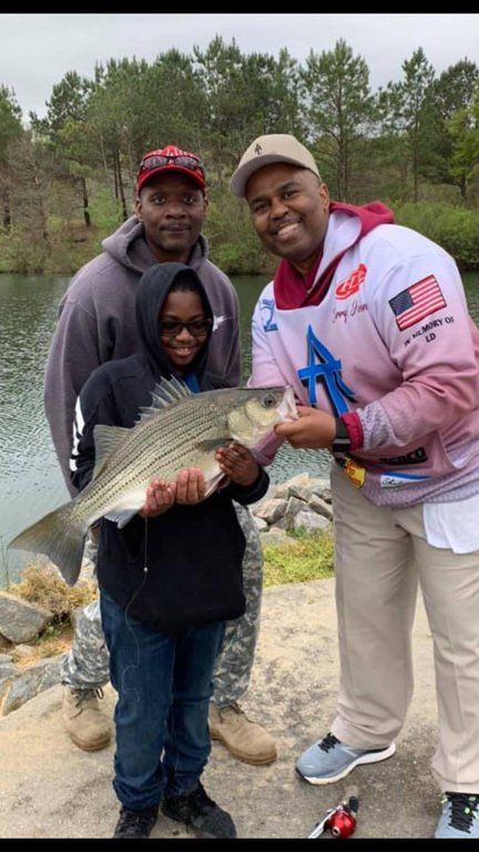 Three people smile, holding a striped bass near a lake.