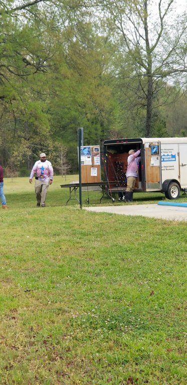 People near a trailer in a grassy park. One man walks toward it, another inside. Trees in the background.