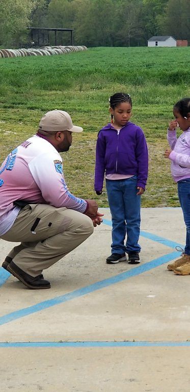 A man in uniform crouches to talk to two young girls on a paved area.