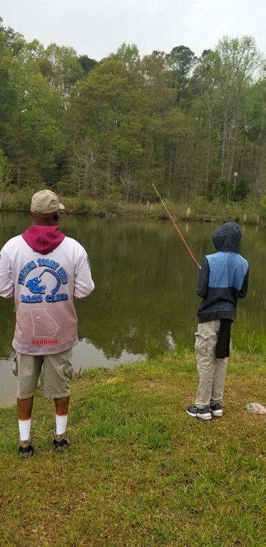 Two people fishing by a pond. An adult wearing white and gray shorts and a child with a blue jacket.