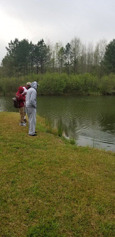 Two people standing near a pond, cloudy day, green grass, trees in background.