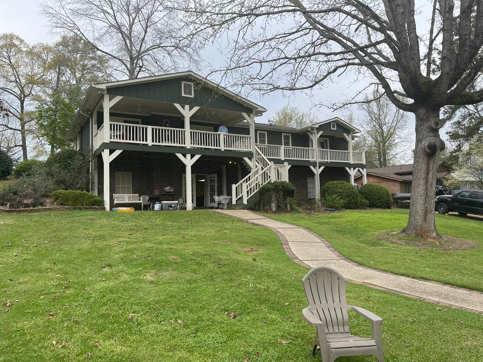 Two-story green house with white balconies, stairs, and pathway in a yard with a chair.