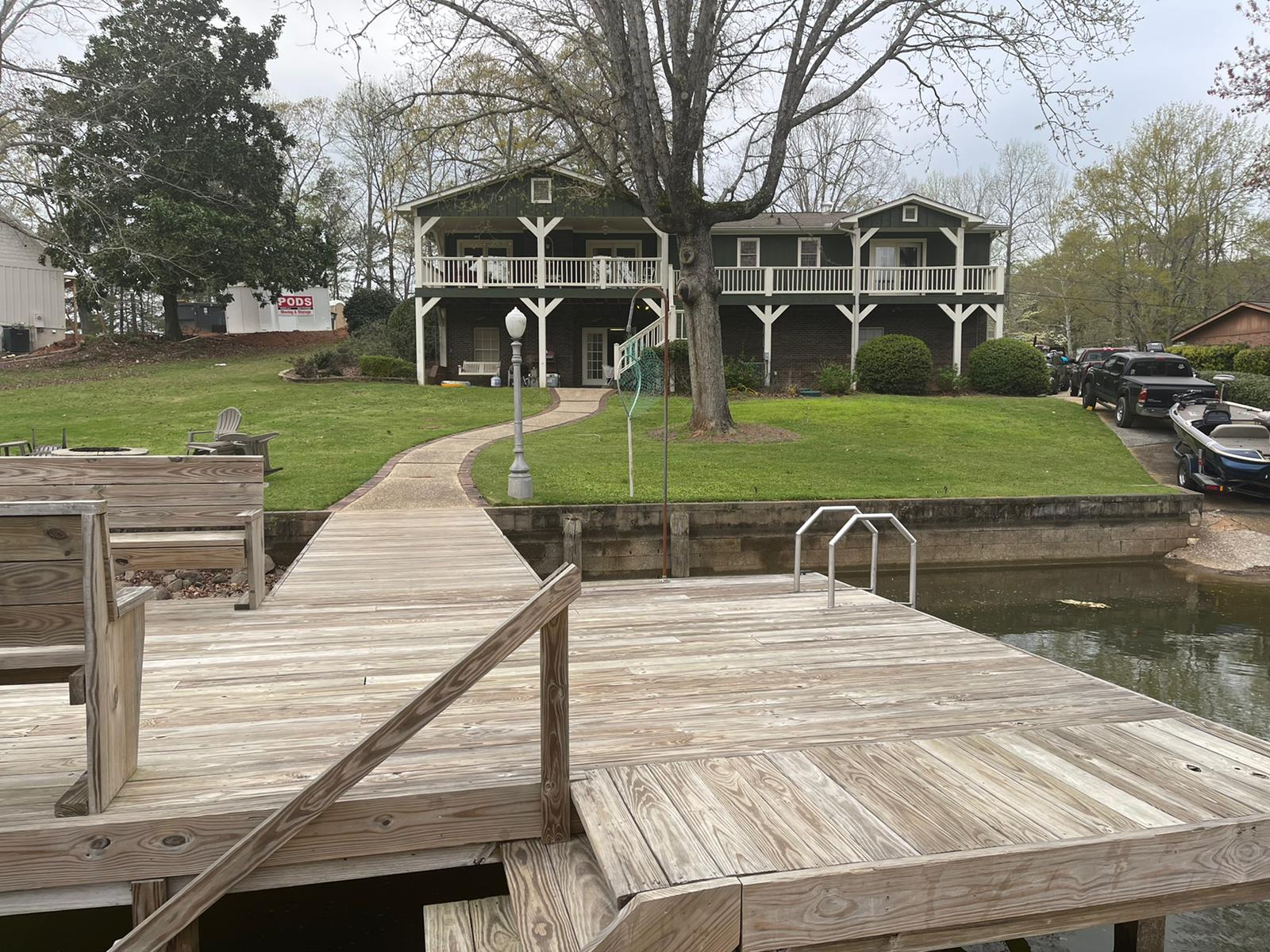 Dock leading to a two-story green house with a balcony, set on a grassy waterfront, overcast sky.