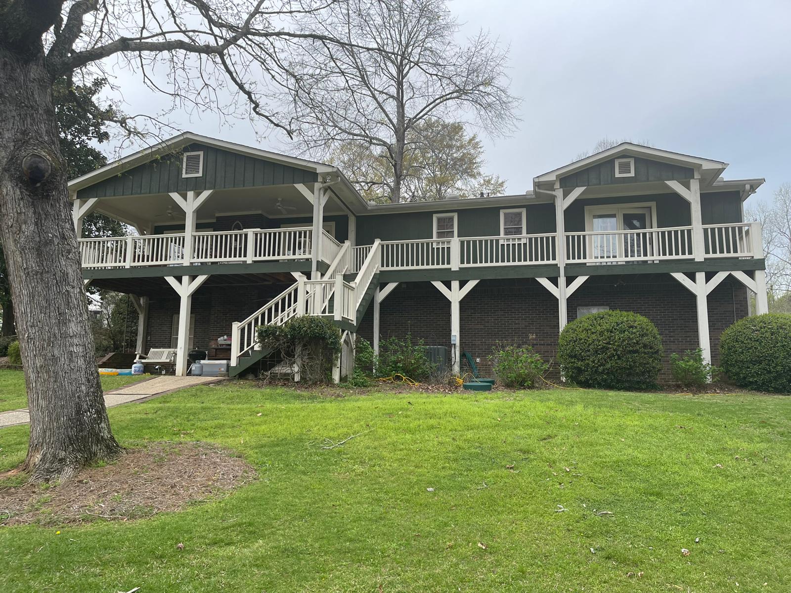 Green two-story house with white railings and porch, on green grass. Cloudy sky.