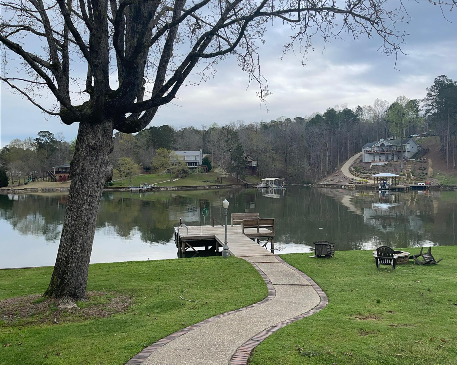 A lake scene with a dock, path, and houses on the opposite shore; overcast skies.