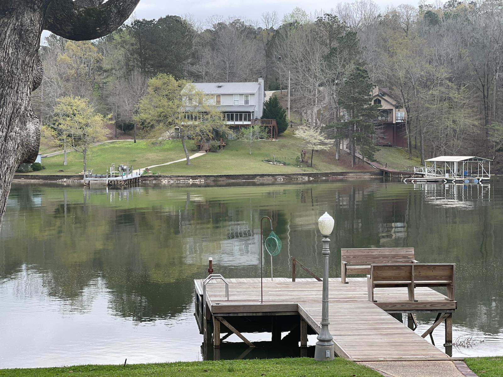 Wooden dock on a lake with a house on a hill in the background, surrounded by trees. Overcast sky.