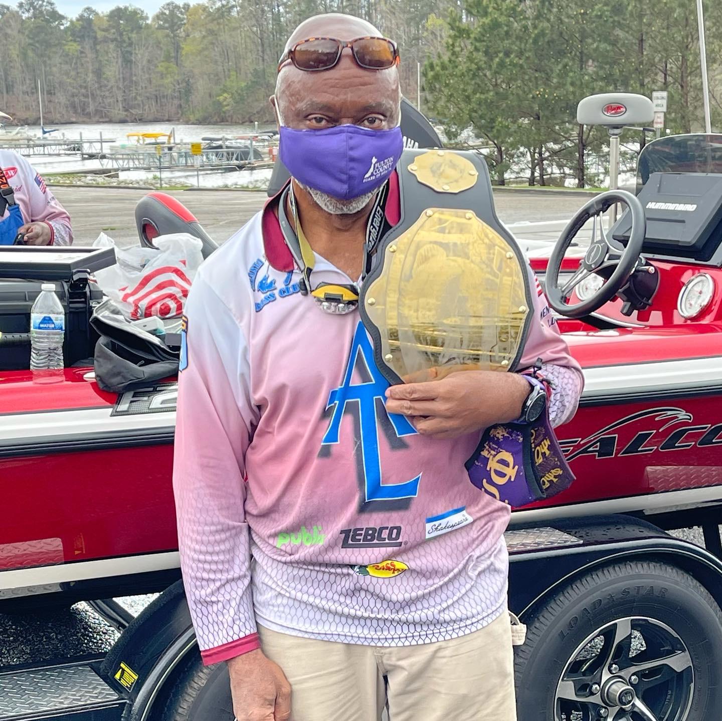 Man holding a gold championship belt with a boat in the background, wearing a mask and sunglasses.