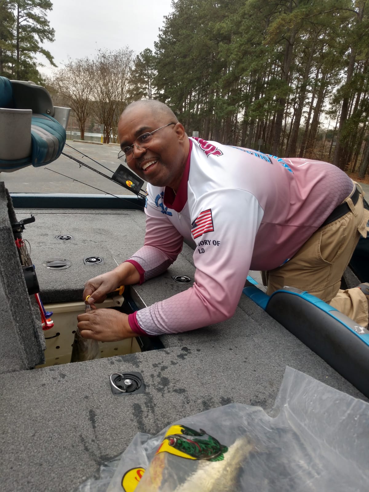 Man in fishing boat, smiling, leaning over compartment, wearing long-sleeved fishing shirt, outdoor setting.