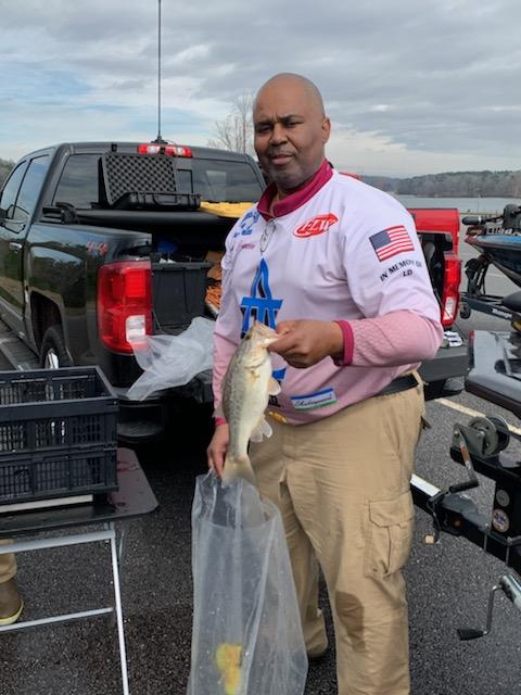 Man holding a fish next to a truck at a boat ramp. He wears a pink and white shirt and tan pants. Cloudy day.