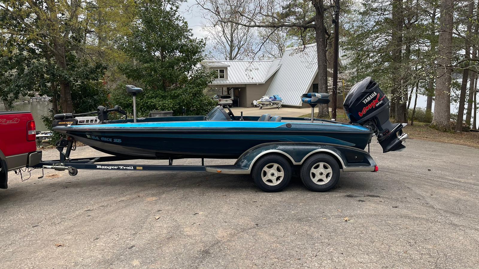 Dark blue bass boat on a trailer, parked on a gravel driveway near a lake, with trees and a house in the background.