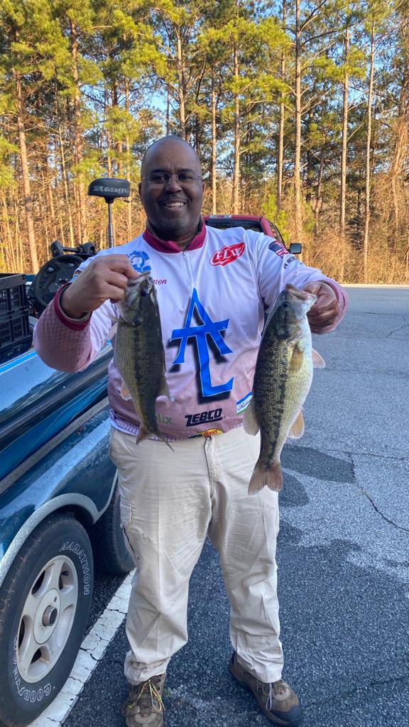 Man in fishing gear holds two bass, smiling near a car outdoors.