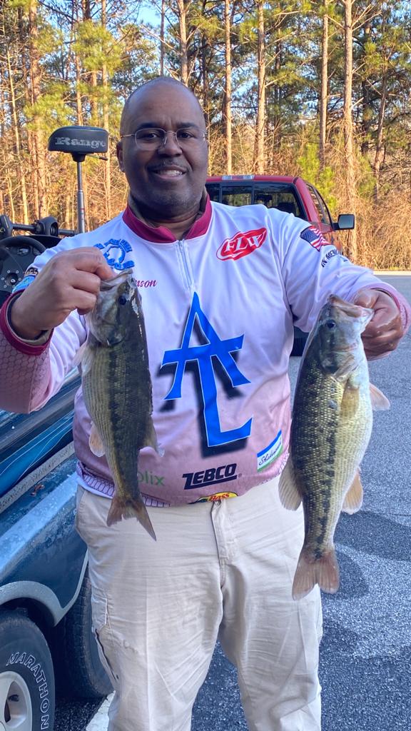 Man holding two bass fish, smiling at the camera near a boat and red truck. He wears fishing apparel.