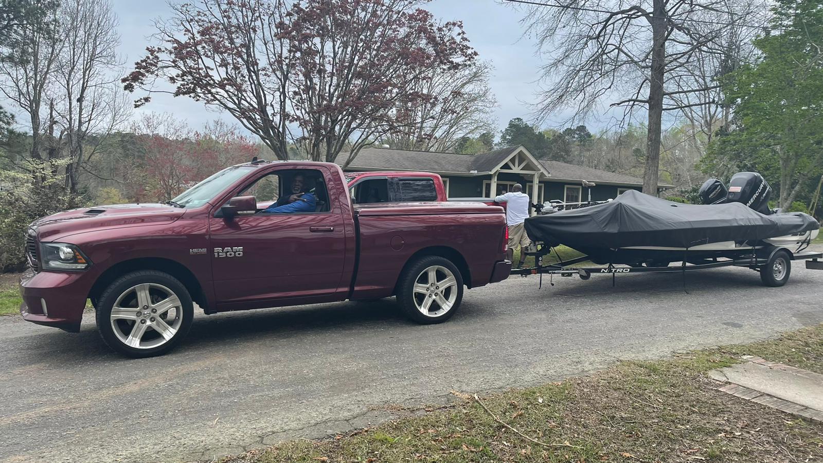 Red pickup truck towing a black boat on a trailer down a driveway.