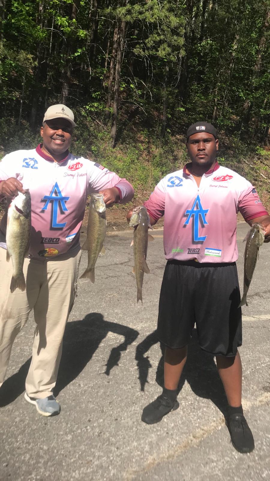 Two men holding up bass they caught, smiling. Outdoors, sunny, wearing fishing shirts and hats.