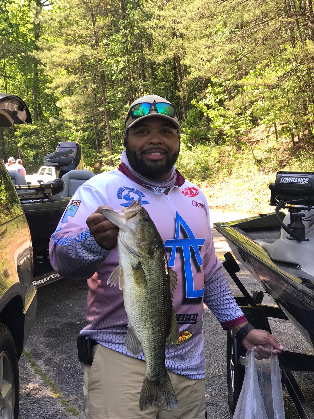 Man in fishing gear holding a largemouth bass near a boat and trees. He’s smiling, wearing sunglasses.