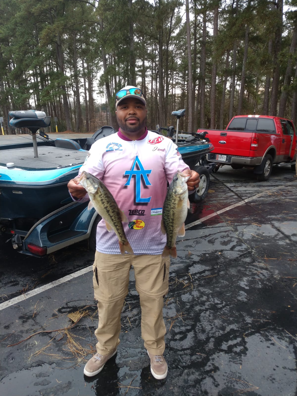 Man holding two bass at a boat dock. He wears a fishing jersey and khaki pants. Red truck in the background.