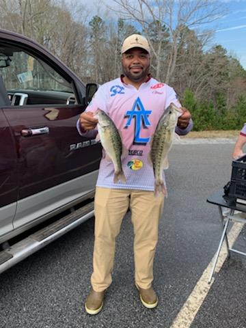 Man holding two bass, standing next to a maroon truck on asphalt, sunny day.