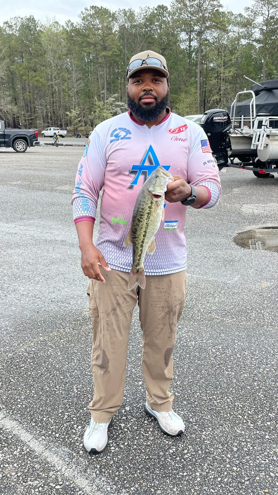 Man in fishing gear holding a bass near boats, wearing a colorful shirt and khaki pants.