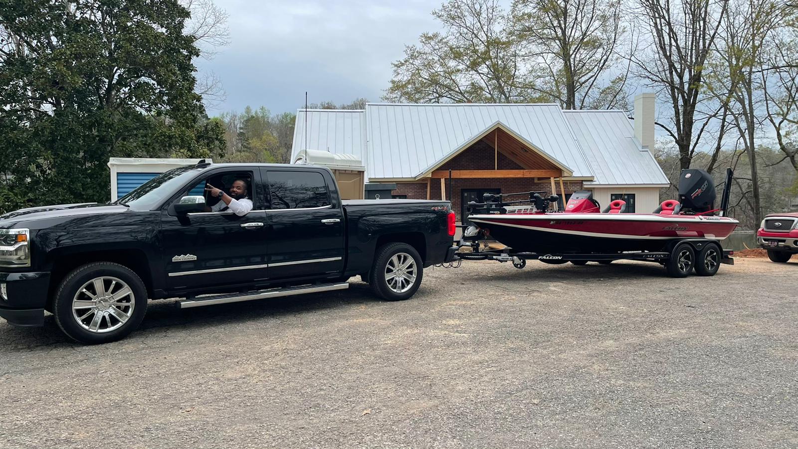 Black truck towing a red boat with a person waving near a white building.