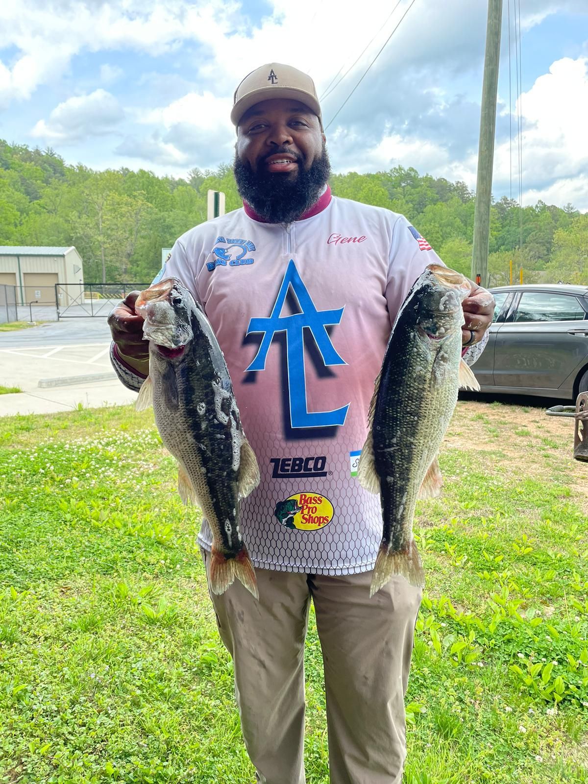 Man holding two fish, smiling. Outdoors with trees, wearing a hat and a logo shirt.