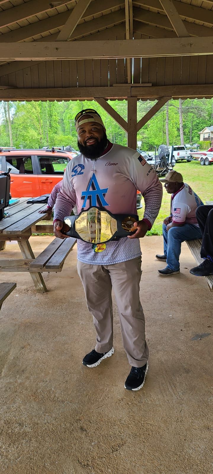 Man holding championship belt under a pavilion; other people and vehicles in the background.