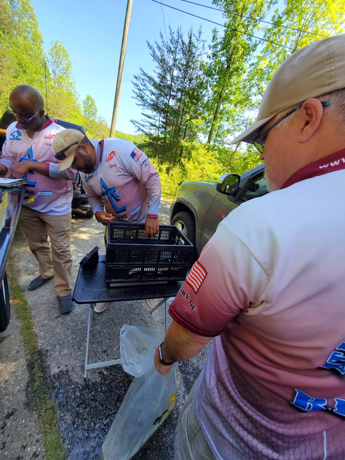 Three men sorting items from a crate outdoors, one holding a clear bag; sunny, wooded area.