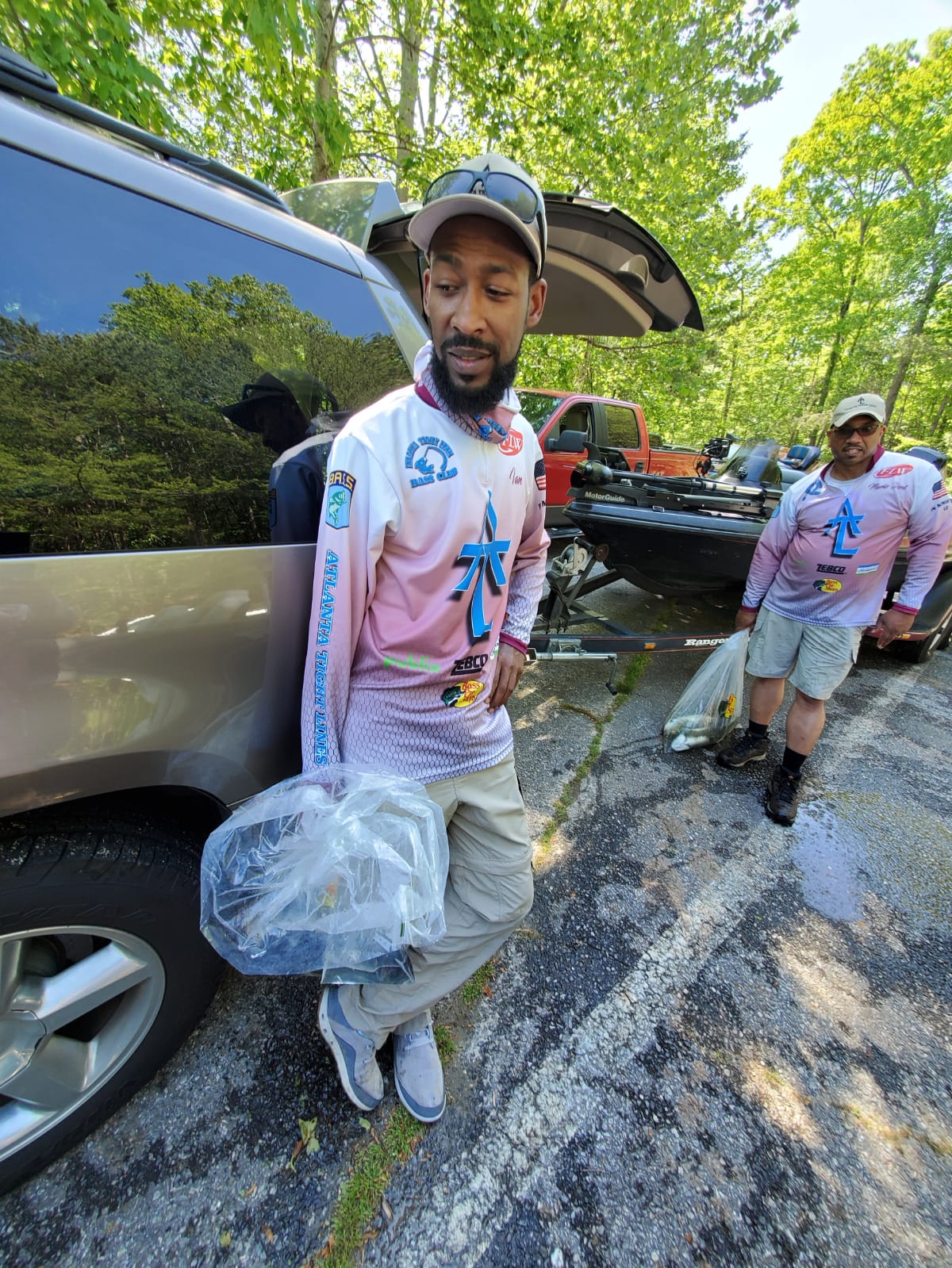 Man in pink shirt holding bag, standing near car and another man outdoors.